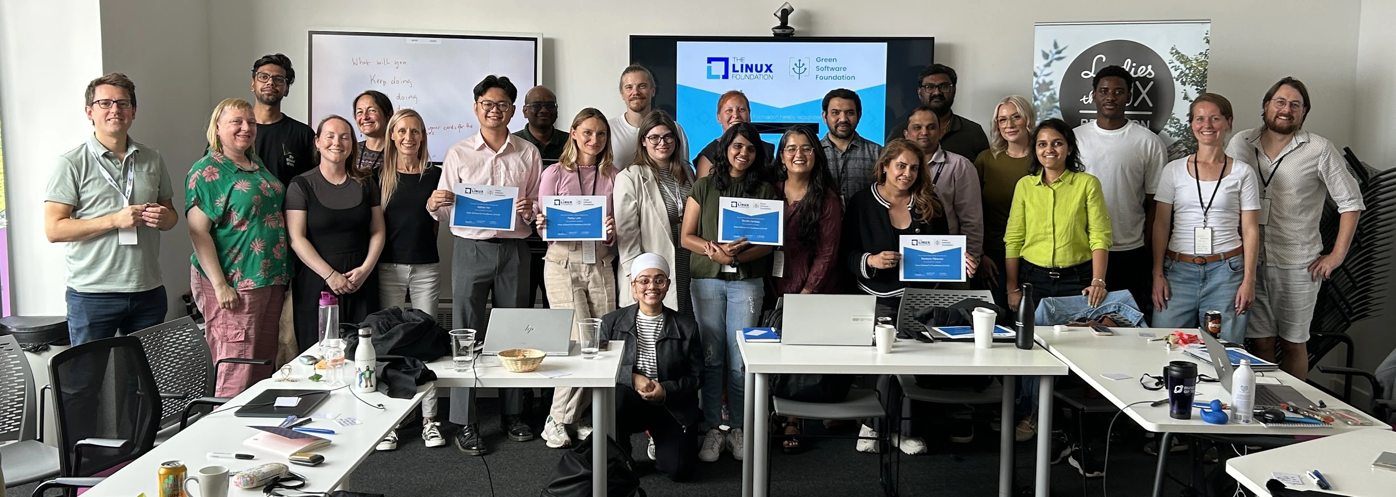 A group people standing in a row in front of an LED monitor with a certificate shown on it. They are smiling and some are holding up their Green Software Practitioner Certificates.