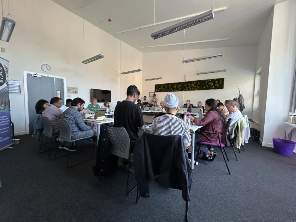 A group of people are sat is a square of tables in the centre of the room, focused on deep study on the Study Day content.