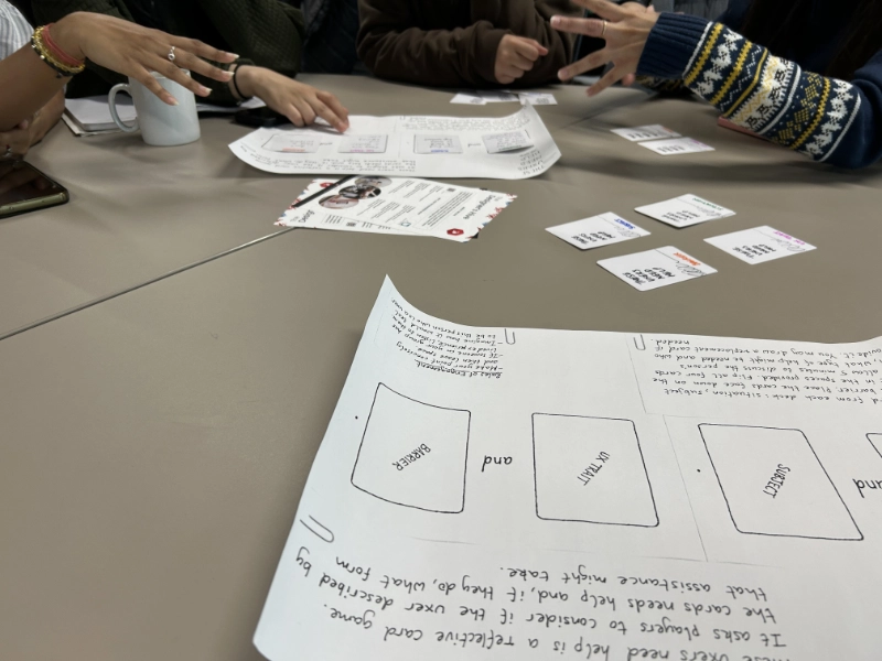 Fiona is smiling while they stand next to a group of students collaborating at a table who are feeding back on their reflections from the 'These UXers Need Help' game